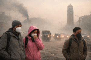 देहरादून की सड़कें घने स्मॉग और प्रदूषण से ढकी हुई हैं, वयस्क लोग मास्क पहनकर धुंध में चल रहे हैं, पीछे क्लॉक टावर और वाहन दिख रहे हैं।